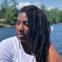 this a photo of Bre. The background is of a lake. Bre is wearing a white shirt and has locs in her hair.